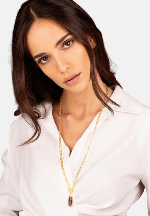 Collana d'oro con pendente a goccia, indossata sopra una camicia bianca. La texture della catena è liscia e il pendente presenta motivi scuri irregolari.