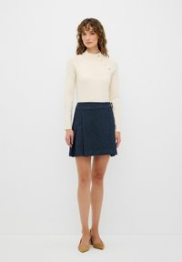 Krämfärgad figurnära tröja med hög krage och fyra dekorativa knappar, tillsammans med en mörkblå plisserad minimini kjol med sidodragkedja.