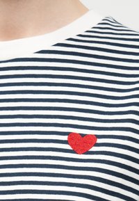 Μπλε ναυτικό και λευκό ριγέ πουκάμισο με μικρή κόκκινη κεντημένη καρδιά στο στήθος κοντά στη λαιμόκοψη.