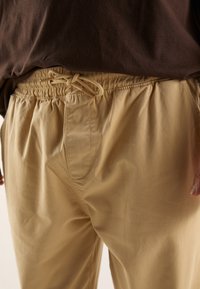 Beżowe spodnie z elastycznym pasem i sznurkiem do wiązania, z bocznymi kieszeniami i miękką, gładką fakturą, częściowo wpuszczone pod brązową koszulę.