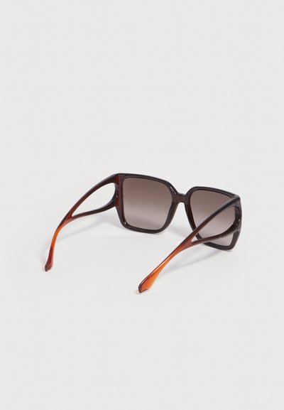 Lunettes de soleil carrées dégradées marron avec des montures épaisses et des branches ouvertes découpées, reposant sur une surface blanche.
