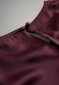 Burgunderrotes Satintop mit einem Rückenschlitzverschluss, der einen runden, mit Stoff bezogenen Knopf und eine glatte, glänzende Textur aufweist.