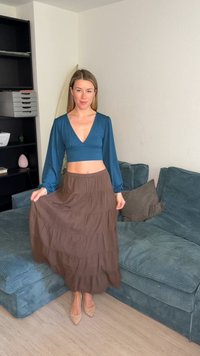 Μια γυναίκα με μπλε crop top μακρυμάνικο και καφέ φούστα με στρώσεις στέκεται κρατώντας τη φούστα της σε ένα σαλόνι με μπλε καναπέ και ράφια πίσω της.