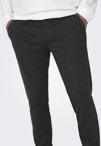 Pantalones ajustados de color gris oscuro hechos de tela suave, con bolsillos frontales y una textura lisa, llevados con una blusa de manga larga blanca.
