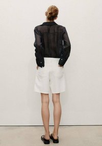Chemise noire transparente à manches longues avec col, associée à un short blanc jusqu'aux genoux. Sandales noires à glissière visibles. Fond uni.