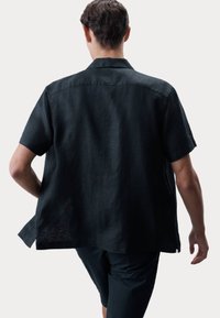 Camisa de lino negra de manga corta con un ajuste holgado, que presenta un yugo trasero y un dobladillo curvado. La tela tiene una apariencia texturizada y ligera.
