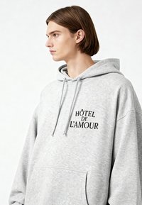 Szürke kapucnis pulóver, kenguruzsebbel és "HÔTEL DE L'AMOUR" fekete felirattal. Lazán oversized fazonú és puha tapintású.