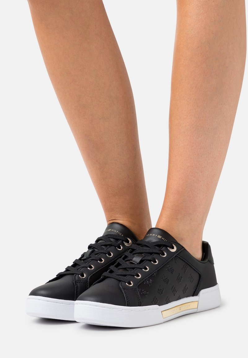 Jambes portant des baskets basses noires avec semelles blanches, lacets noirs et un marquage subtil à motifs sur les côtés, sur fond blanc.