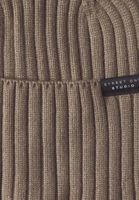 Gorra de punto marrón con textura de canalé vertical. Presenta una etiqueta negra con texto blanco que dice "STREET ONE STUDIO" cosida en el lateral.