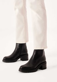 Stivaletti neri in pelle con tacco spesso, dotati di cerniera laterale, suola a coste e design elegante, abbinati a pantaloni bianchi.