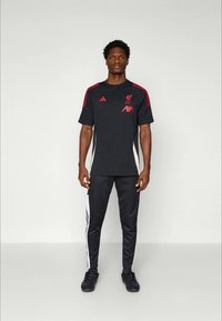 Must spordisärk punaste detailidega, millel on logo ja tekst 'AXA'. Sobitatud mustade pükstega, mille külgedel on valged triibud. Mustad tossud.