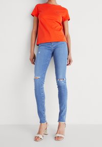 Camiseta de algodón naranja con mangas cortas combinada con jeans ajustados de un tono azul claro que presentan detalles rasgados y un ligero desgaste, complementada con sandalias de tacón bloque en color beige.