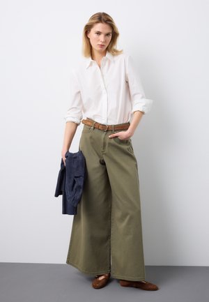 Femme portant une chemise blanche boutonnée, un pantalon large vert olive avec ceinture marron, tenant une veste bleu marine dans la main gauche, debout contre un mur blanc.