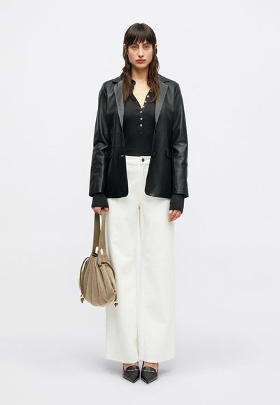 Žena v černé kožené blejzru, černém knoflíkovém topu, bílých širokých kalhotách, černých špičatých botách, držící béžovou kabelku na jednoduchém pozadí.