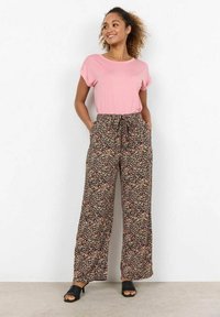 Svetle růžové tričko s krátkým rukávem v kombinaci s černými kalhotami s širokým střihem, které mají květinový vzor v růžové, žluté a bílé barvě. Černé sandály s otevřenou špičkou.