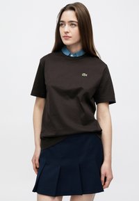 Pruun puuvillane t-särk lühikeste varrukatega, millel on väike roheline Lacoste'i logo. Kandes seda helesinise kraega särgi peal ja ühendades meresinise volditud seelikuga.