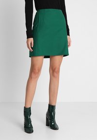 Mini-jupe verte avec une silhouette ajustée, texture lisse et coutures latérales discrètes, associée à des bottines vertes brillantes.