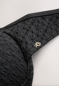 Coque de soutien-gorge noire texturée avec de petites perforations et un petit charm décoratif rond doré sur fond clair.