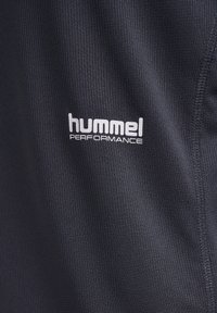 Close-up van zwart gestructureerd stof met wit "hummel PERFORMANCE"-logo en zichtbare stiksels langs de rechterrand.