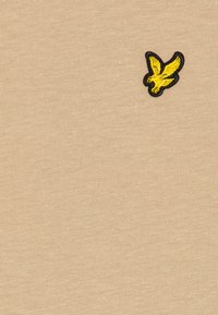 Tissu en coton beige avec un petit logo d'oiseau brodé noir et jaune sur le coin supérieur gauche. Texture lisse et tissage régulier.