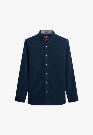 Chemise en velours côtelé bleu marine avec col boutonné, manches longues et un logo rouge sur la poitrine. Elle présente des boutons blancs et un tissu texturé.