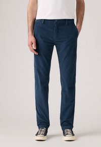 Bărbat îmbrăcat în pantaloni de catifea bleumarin, tricou alb și adidași negri cu tălpi albe, stând în fața unui fundal simplu.