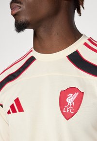 Creme trøje med røde og sorte accenter. Har et Liverpool FC-logo og Adidas-branding på venstre side. Glat stofstruktur.