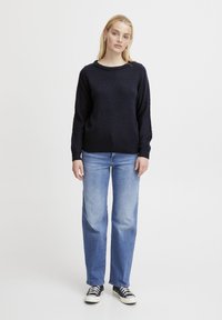 Marineblau gestrickter Pullover mit strukturiertem Muster, rundem Halsausschnitt und geripptem Saum. Kombiniert mit hellblauen Jeans im geraden Schnitt und schwarzen Sneakers.
