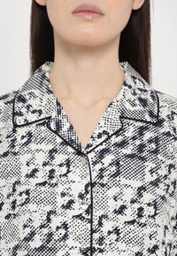 Cămașă albă cu imprimeu abstract negru, având guler, mâneci scurte și închidere cu butoane la față. Textură asemănătoare satinului, cu margini contrastante.