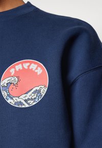 Tengerészkék pulóver, kerek grafikával, amely hullámokat és szöveget ábrázol korall háttéren. Az anyag simának tűnik, bordás mandzsettàkkal.