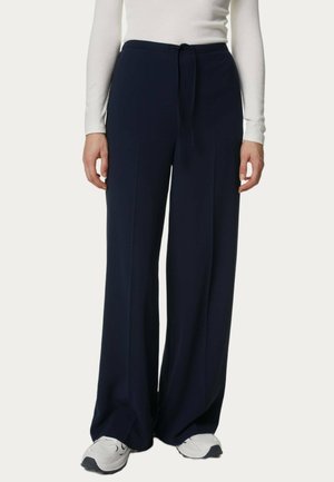 Pantaloni a gamba larga blu marina realizzati in morbido tessuto con vita elasticizzata e pieghe frontali, abbinati a una maglietta bianca a maniche lunghe e sneakers grigie.