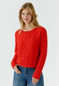 Roter Strickpullover mit strukturiertem Wellenmuster, weitem Ausschnitt und langen Ärmeln, kombiniert mit blauen Jeans.