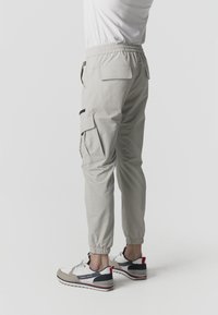 Persona con pantalones cargo beige claro con puños elásticos y una camisa blanca, combinados con zapatillas beige y negras con detalles en rojo.