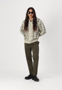 Strikket cardigan i krem med svart rutestruktur, knappestolpe, ribbestrikket kant. Olivengrønne bukser og svarte sko fullfører antrekket.