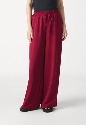 Femme portant un pantalon large ample, taille haute, rouge foncé, avec un cordon à la taille, un haut noir et des sandales plateformes noires.