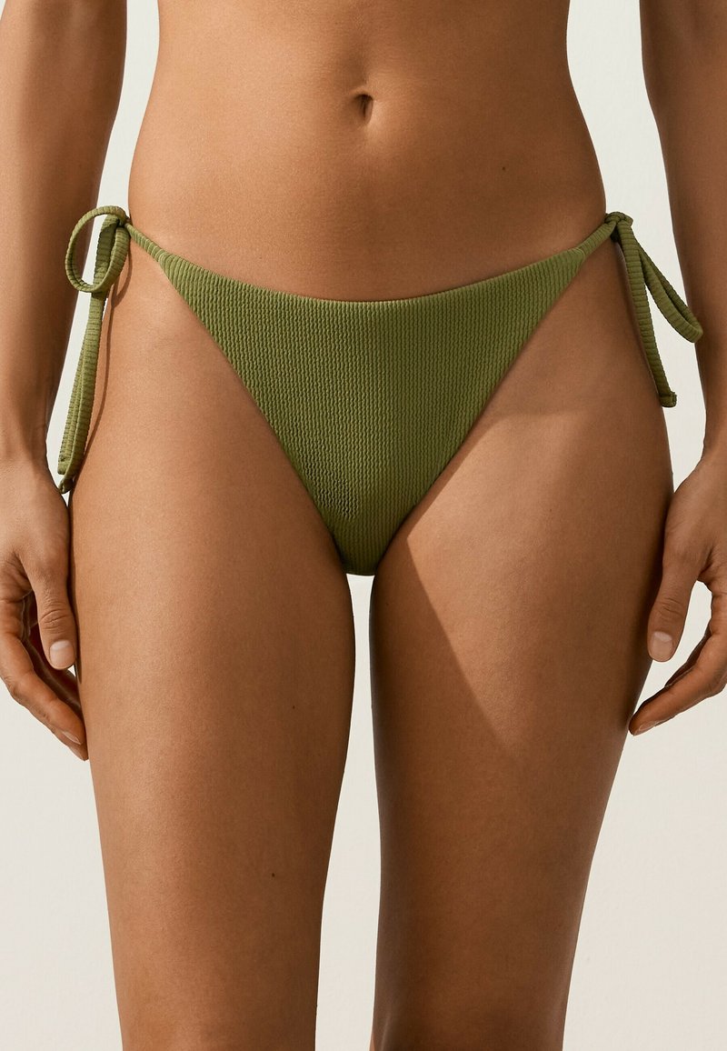 Gros plan d'une personne portant un bas de bikini en tissu texturé vert olive avec des liens latéraux sur un fond neutre.