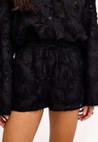 Blouse noire transparente texturée à manches longues et shorts assortis avec taille élastique et cordon de serrage, portés par une personne à la peau marron clair.