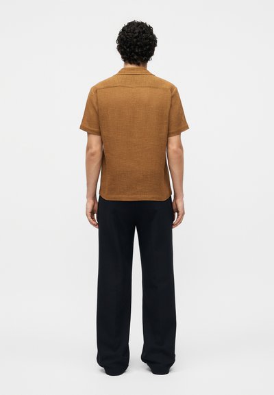 Άνδρας με σγουρά μαλλιά στέκεται με την πλάτη γυρισμένη, φορόντας ένα καφέ σκληρό υφασμάτινο κοντομάνικο πουκάμισο και φαρδιά μαύρη παντελόνια σε απλή φόντο.