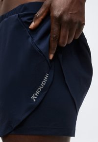 Σκούρο δέρμα χέρι που τραβάει μπλε σορτς Houdini, εστιάζοντας στην πλαϊνή τσέπη και το λογότυπο της μάρκας σε λευκό φόντο.