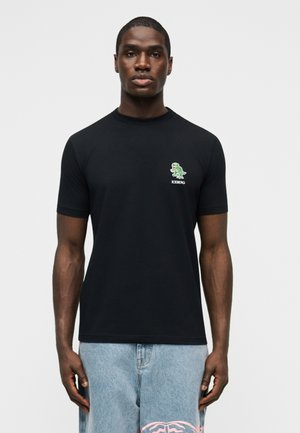 Άνδρας που φοράει μαύρο μπλουζάκι με ένα μικρό πράσινο λογότυπο δεινοσαύρου και το κείμενο «ICEBERG» στο στήθος, συνδυασμένο με ανοιχτόχρωμο τζιν παντελόνι.