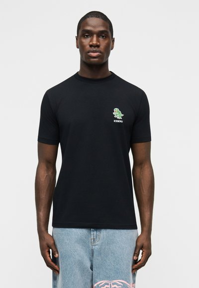 Homme portant un t-shirt noir avec un petit logo de dinosaure vert et le texte "ICEBERG" sur la poitrine, associé à un jean bleu clair.
