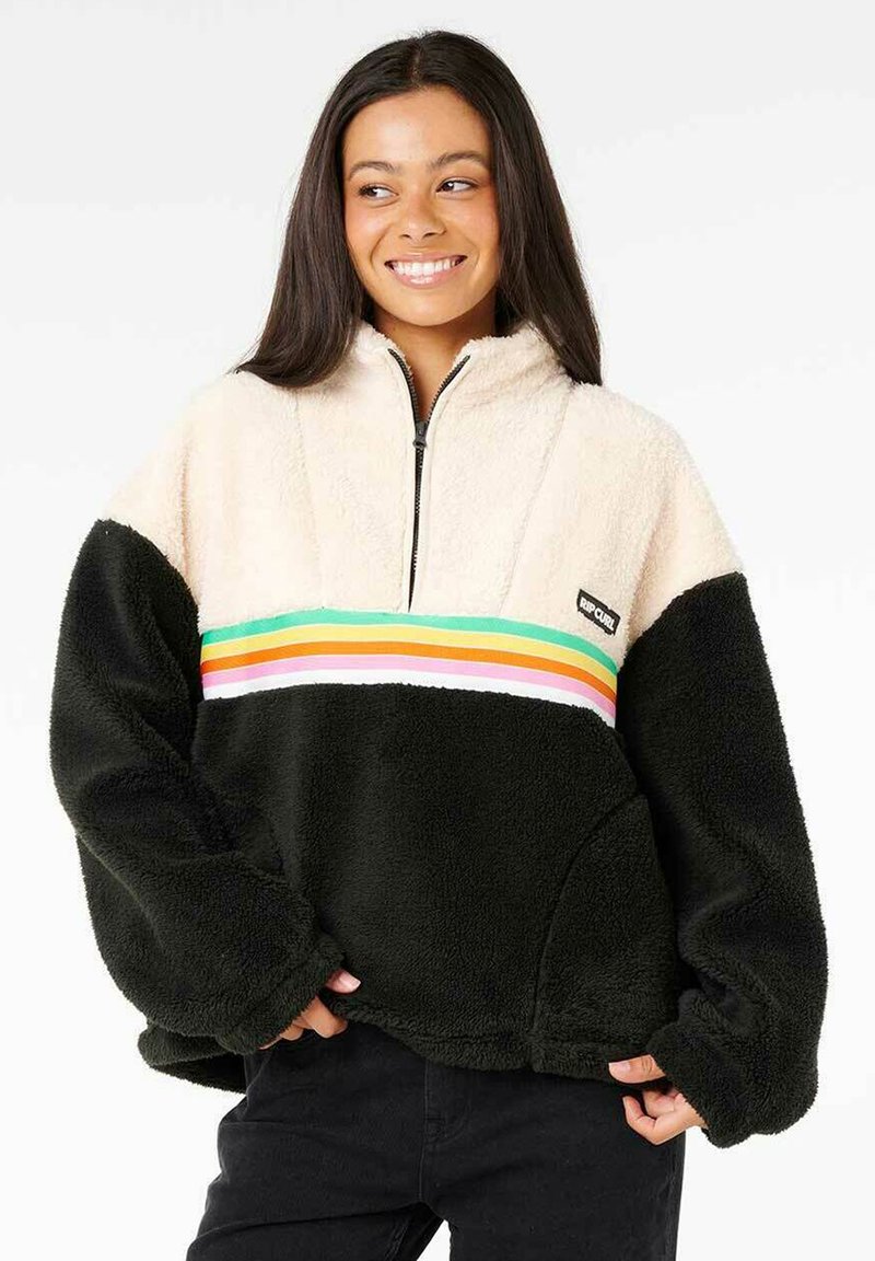 Flauschiger Pullover mit creamfarbener Oberseite und schwarzem Unterteil, der ein mehrfarbig gestreiftes Band über die Brust und einen Reißverschlusskragen hat.