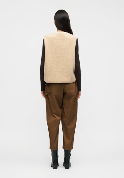 Μπεζ φλις γιλέκο με στρογγυλεμένο τελείωμα, φορεμένο πάνω από ένα σκούρο μακρυμάνικο πουκάμισο, συνδυασμένο με καφέ παντελόνι με στενή γραμμή και μαύρες μπότες με τακούνι.