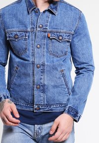 Džínová bunda v světle modré barvě s klasickým střihem, obsahující dvě náprsní kapsy, zapínání na knoflíky a hnědé detaily. Přítomná opotřebovaná textura.