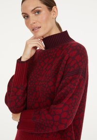 Frau mit heller Haut und zurückgegebendem Haar, die einen rot und dunkelrot gemusterten Rollkragenpullover trägt und nachdenklich ihr Kinn hält.