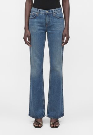 Άτομο που φορά μεσαίας μέσης, μεσαίου μπλε χρώματος καμπάνες τζιν και μαύρα ανοικτά σανδάλια με χρυσή αλυσίδα, στέκεται μπροστά σε λευκό φόντο.