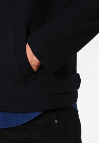 Dunkelblaue Jacke mit glatter Textur, ausgestattet mit einer Seitentasche und einem gefalteten Detail am Saum. Wird über einem blauen Hemd getragen.