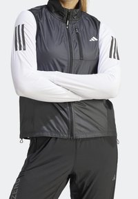 Czarna i szara sportowa kamizelka z wysokim kołnierzem, zamkiem błyskawicznym oraz trzema paskami na białych rękawach; wykonana z gładkiego, lekkiego materiału.