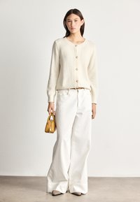 Crèmekleurige cardigan met een structuurtje en vijf knopen, gecombineerd met witte rechte jeans. Houdt een kleine gele handtas vast. Vierkante, beige schoenen.