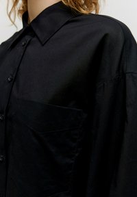 Schwarzes Hemd mit Knöpfen, aus glattem Stoff, mit Kragen, einer Brusttasche und dezenten Nähdetails.